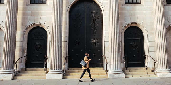 A woman walks in the street looking at her phone. There are large doors in the background of the image on an old building.