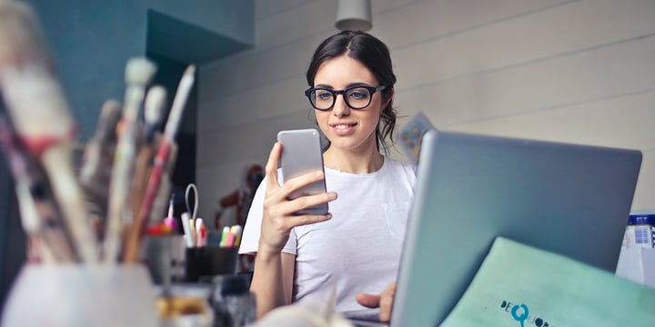 Caucasian woman wearing glasses holding her phone up and looking at it. She is in a home study environment.