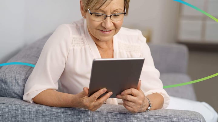 Aileen, 64, sitting on a sofa holding her tablet device and smiling.