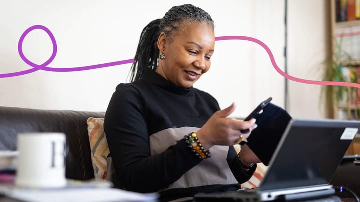  A 55 year old black lady sitting on the sofa holding her phone and smiling.