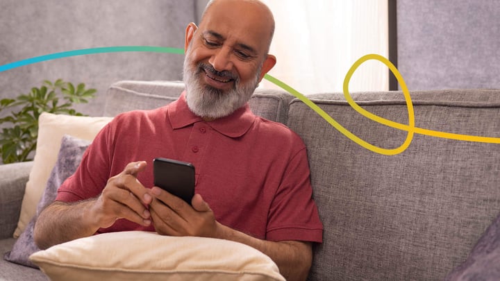 55 year old Asian man sitting on the sofa and using her mobile phone whilst smiling.