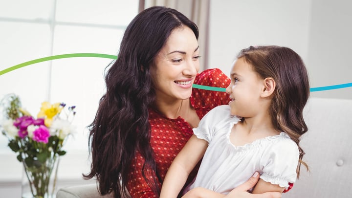 A Caucasion woman with black hair is sitting on a sofa with her young daughter on her lap. They are looking at each other whilst smiling,
