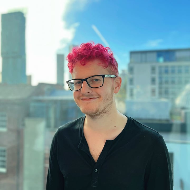 A person with bright pink hair smiling, with a city view in the background.