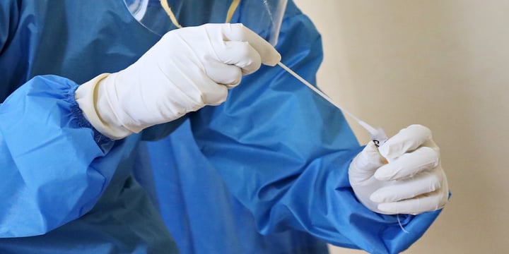 Image of a nurse putting a swab in a test tube wearing latex gloves. Only the hands and swab is imaged with a blue uniform shown in the background.