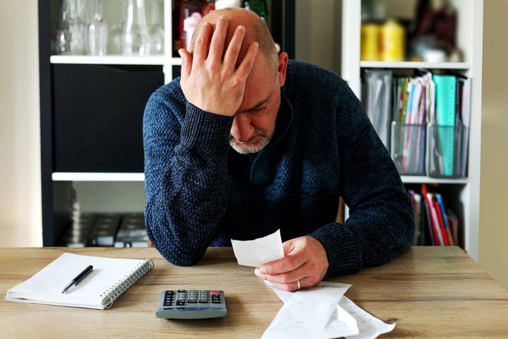 Distressed man sitting at a table looking at bills with his hand on his head.