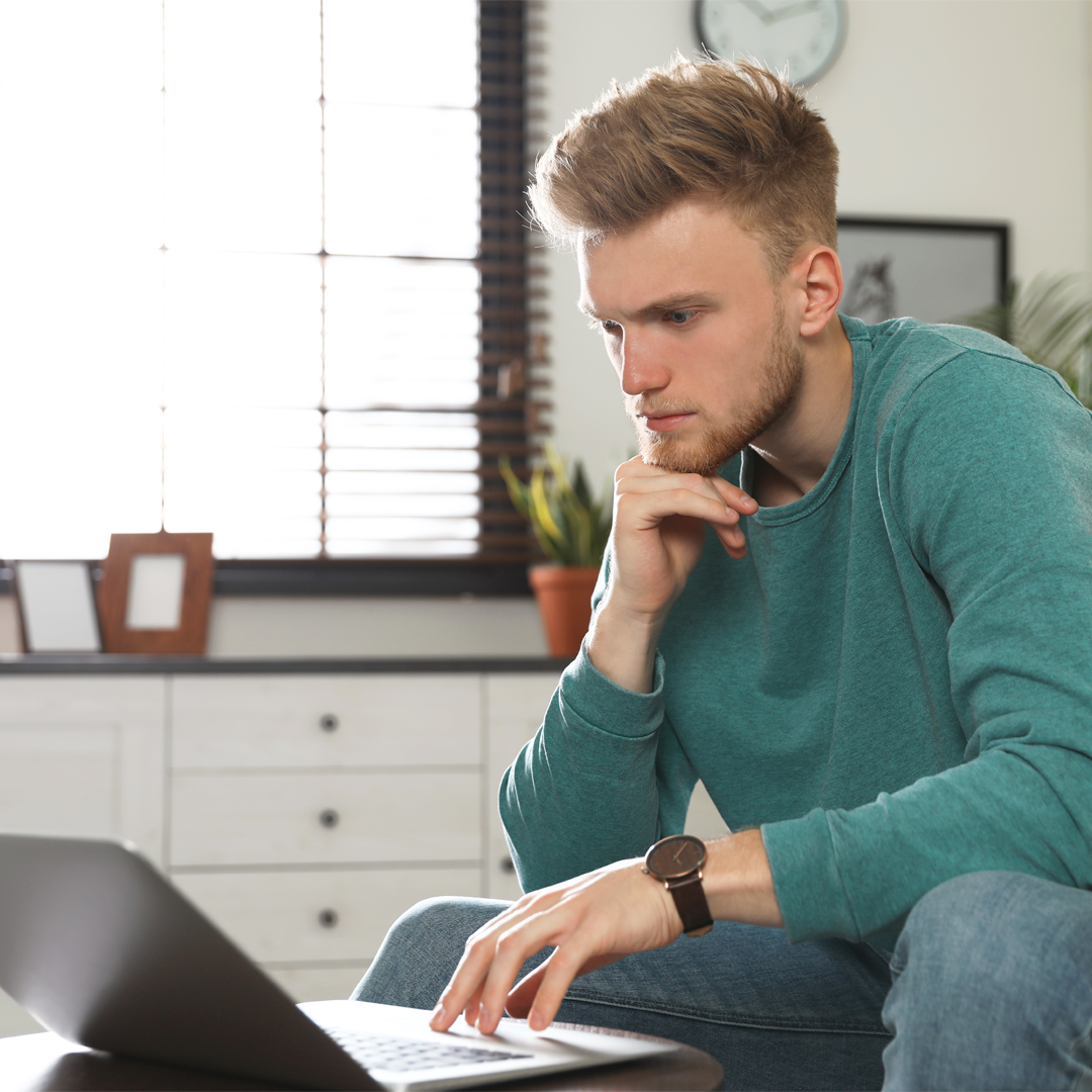 young male looking at his laptop whilst sitting on the sofa 1080 x 1080 px